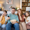 Familie sitzt gemeinsam auf dem Sofa, schaut einen Film über Geld und isst dabei Popcorn.