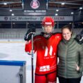 Junge in Eishockeyausrüstung steht in einer Eishalle, daneben steht seine Mutter in einer Winterjacke.